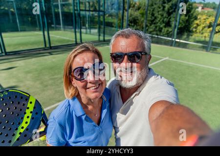 Das Senior Padel-Paar, beide mit Sonnenbrille, feiert ihren Sieg mit einem Selfie auf dem Platz. Lächelnd und stolz fangen sie die fröhliche Mutter ein Stockfoto