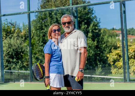 Ein älteres Padel-Paar mit Sonnenbrille posiert eng auf dem Platz, lächelt und hält die Paddel. Mit Stolz und Freude umarmen sie die M Stockfoto