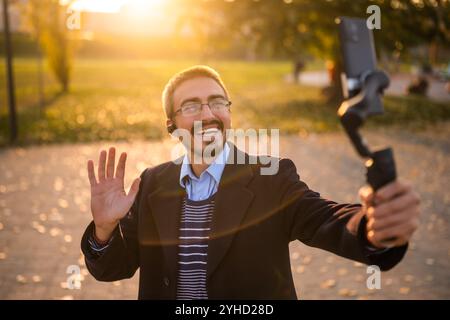 Geschäftsmann-Vlogger, der Ohrhörer trägt und winkt, während er ein Mobiltelefon mit Gimbal benutzt. Er steht im Park und nimmt Vlog auf. Stockfoto