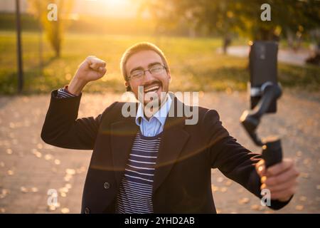 Geschäftsmann-Vlogger, der Ohrhörer trägt und ein Mobiltelefon mit Gimbal zur Aufzeichnung von Vlog verwendet, während er an einem sonnigen Herbstnachmittag im Park steht. Stockfoto