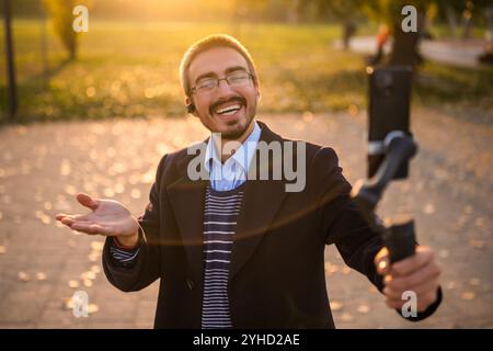 Geschäftsmann-Vlogger, der Ohrhörer trägt und ein Mobiltelefon mit Gimbal zur Aufzeichnung von Vlog verwendet, während er an einem sonnigen Herbstnachmittag im Park steht. Stockfoto