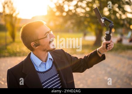 Geschäftsmann-Vlogger, der Ohrhörer trägt und ein Mobiltelefon mit Gimbal zur Aufzeichnung von Vlog verwendet, während er an einem sonnigen Herbstnachmittag im Park steht. Stockfoto