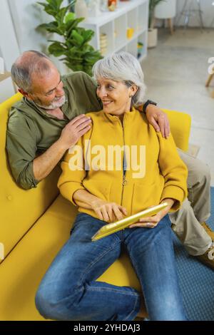 Vertikales Foto eines entspannten Seniorenpaares, das mit einem digitalen Tablet auf dem Sofa liegt Stockfoto