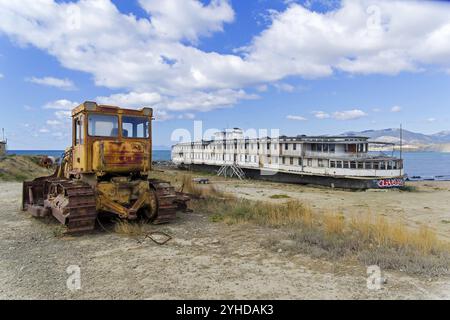 Ein altes verlassenes Flussboot steht am Boden neben einem rostigen Bulldozer in der Nähe des Meeres. Bucht Kapsel, Krim. Sonniger Tag im September Stockfoto