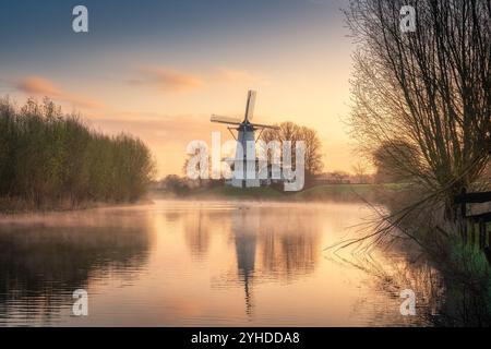 Friedlicher Sonnenaufgang über einem Nebelfluss mit traditioneller niederländischer Windmühle in den Niederlanden Stockfoto