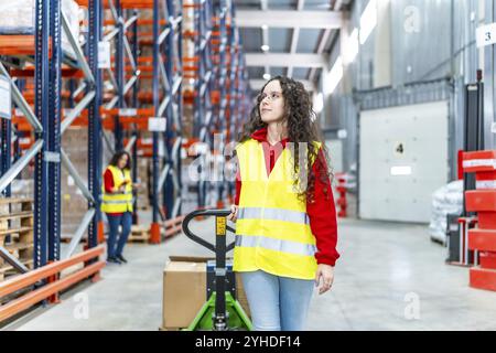 Junge kaukasische Arbeiterin, die einen Wagen voller Kisten schiebt, die einen Verteilerlagerkorridor entlang laufen Stockfoto
