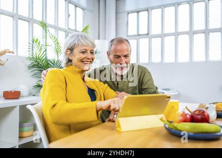 Glückliches Seniorenpaar, das zu Hause ein digitales Tablet verwendet, auf den Bildschirm zeigt und lächelt, während es zusammen frühstückt Stockfoto