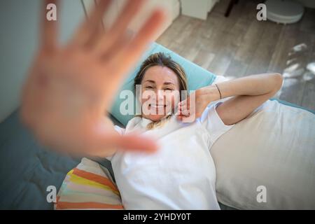Eine verschlafene Frau mittleren Alters liegt auf dem Bett, lächelt und dehnt sich mit einer Hand nahe an der Kamera. Stockfoto