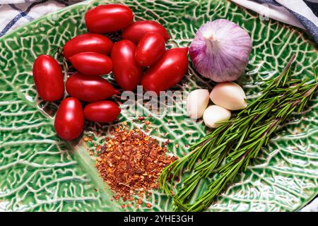 Reife Pflaumentomaten, Rosmarin, Knoblauch und Chilischoten auf der grünen Platte mit Blattmuster Stockfoto