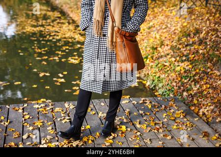 Frau mit Hahnentrittmantel, Kaschmirschal und Ledertasche steht am See im Herbstpark. Trendige Mode warme Kleidung Stockfoto