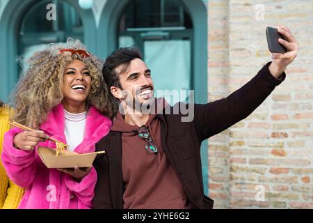 Multiethnische Freunde teilen lustige Selfie-Momente, während sie asiatische Speisen zum Mitnehmen in der City Street genießen Stockfoto