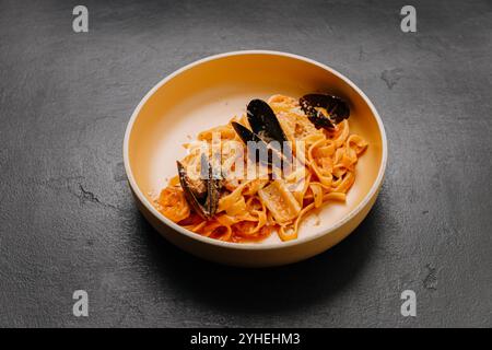 Tagliatelle-Pasta mit Muscheln und reichhaltiger Tomatensauce in einer beigen Schüssel auf dunklem, strukturiertem Hintergrund Stockfoto