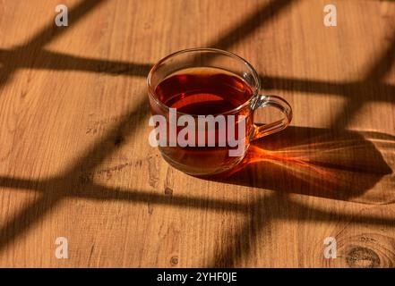 Eine zarte Glasschale mit Bernsteintee liegt auf einer rustikalen Holzoberfläche, die von sanftem Nachmittagslicht beleuchtet wird. Stockfoto
