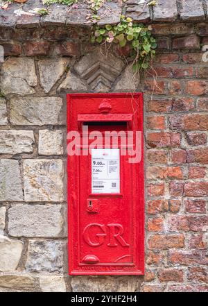 Ein historischer roter Postkasten, der an einer Ziegelsteinmauer befestigt ist. Blätter einer kriechenden Pflanze kommen über die Wand und hängen herunter. Postinformationen Stockfoto