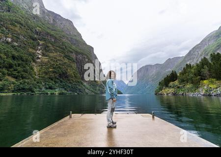 Eine junge Frau, um 30 Jahre alt, steht an einem bewölkten Tag auf einem kleinen Dock und blickt auf die Kamera vor dem Hintergrund der weiten Landschaft des Nærøyfjord. Die Stockfoto