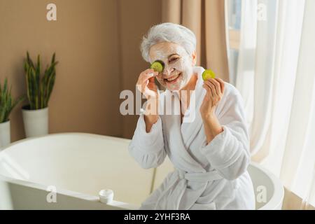 Reife Erwachsene alte Frau mit pflegender Anti-Age-Gesichtsmaske, die Gurkenscheiben hält. Ältere Frau mit weißer Maskencreme auf der Haut bekommt Gesichtsbehandlung tägliche Schönheitsroutine. Hautpflege Alterserscheinung Stockfoto