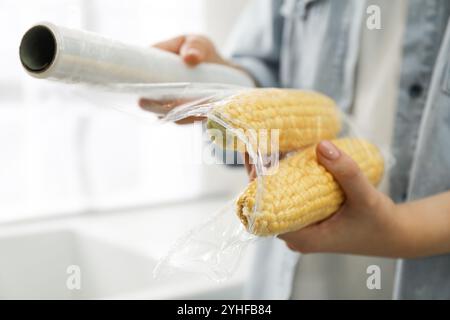 Frau, die Plastikfutterverpackung über Maiskolben in der Küche legt, Nahaufnahme Stockfoto