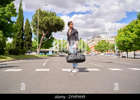 Indischer Mann in schwarzem Hemd und zerrissener Jeans, der auf einem Straßenübergang steht und eine schwarze Tasche mit Bäumen und Gebäuden im Hintergrund hält. Stockfoto