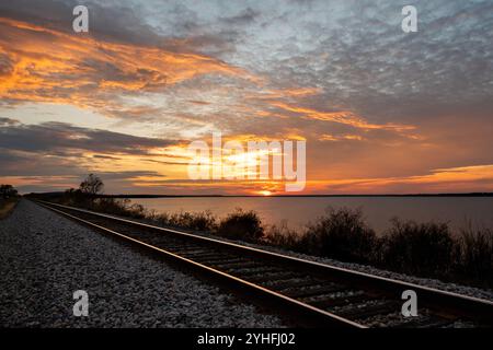 Eisenbahngleise führen in die Ferne entlang des Ufers eines Sees, während der Sonnenuntergang den Himmel mit feurigen gelben und orangen Wolken erfüllt. Stockfoto