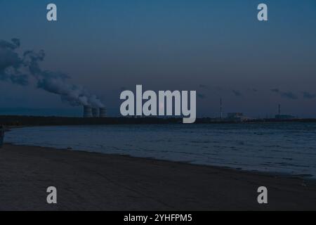 Industrieküste in der Abenddämmerung mit Rauchstapeln, die Rauch in den Himmel ausstoßen, von einem Strand aus gesehen. Ruhiges Wasser und ferne Horizonte erzeugen eine stimmungsvolle Atmosphäre Stockfoto