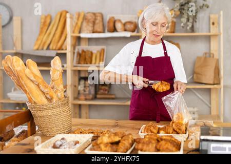 Die Senior-Verkäuferin bringt Croissant in eine Plastiktüte für den Käufer, verpackt Waren. Stockfoto