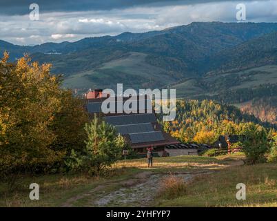 Ein malerischer Blick auf eine Berghütte mit Solarpaneelen, umgeben von sanften grünen Hügeln und Herbstlaub. Ein Wanderer nähert sich dem Tierheim nach einsam Stockfoto