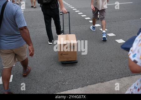 Ein Mann zieht einen Pappkarton auf einem Trolley über eine Straßenkreuzung in Singapur. Blickpunkt von einer ersten Person, nachdem sie auf die Füße geschaut hat. Stockfoto