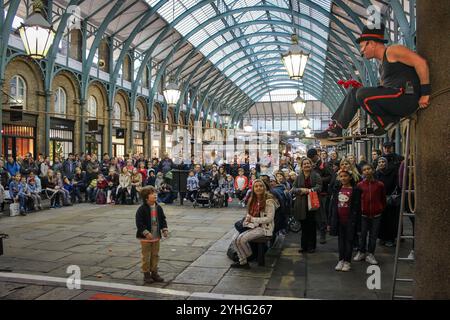 Ein Straßenkünstler unterhält eine Menge in einer Markthalle, während ein Kind den Künstler ansieht, während das Publikum amüsant zusieht. Stockfoto