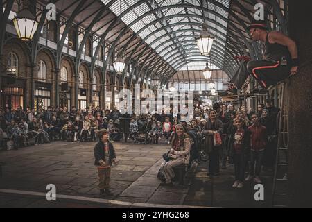 Eine Menschenmenge versammelt sich in einer geschäftigen Marktstraße, beobachtet einen Künstler, der eine Säule klettert und mit seinen Acts unterhält. Stockfoto