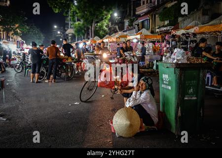 Eine lebhafte Nachtmarktszene in Hanoi, auf der sich die Einheimischen treffen, um unter dem Licht Obst und Waren zu verkaufen, während zwei ältere Frauen eine kurze Pause einlegen. Stockfoto