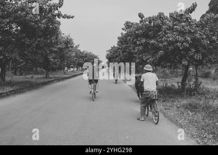 Ein Schwarzweiß-Foto zeigt zwei Männer, die auf einer ruhigen, von Bäumen gesäumten Landstraße Fahrrad fahren. Stockfoto