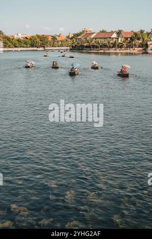 Boote voller Touristen paddeln entlang eines friedlichen Flusses mit traditionellen vietnamesischen Häusern und Palmen, die die Ufer unter klarem Himmel säumen. Stockfoto