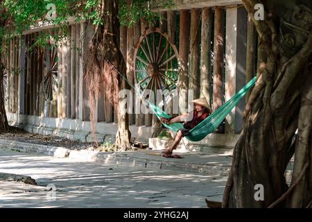 Ein entspannter, älterer vietnamesischer Mann, der in einer grünen Hängematte unter strahlendem Sonnenlicht ruht und einen traditionellen konischen Hut trägt, umgeben von einer rustikalen Kulisse Stockfoto