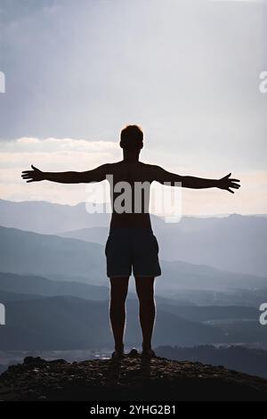 Silhouette eines Mannes mit offenen Armen, der auf einem Berggipfel steht und die Weite der Natur umhüllt, mit Schichten von Bergen, die sich bis zum Horizont erstrecken. Stockfoto