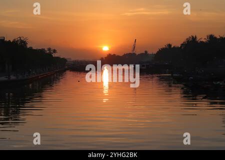 Sonnenaufgang über einem ruhigen Fluss in Hoi an, Vietnam, während die Sonne ein warmes Spiegelbild auf das Wasser und Silhouetten von Bäumen und Booten wirft. Stockfoto