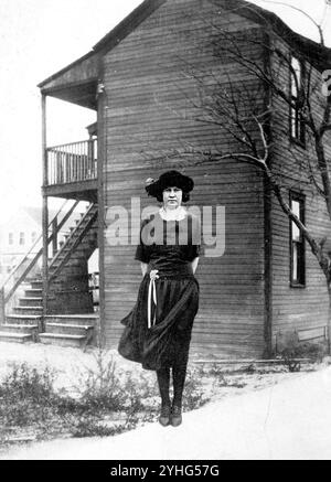 Authentische Vintage-Fotografie aus den 1920er Jahren einer Frau in modischem Kleid und Hut, die vor dem Holzhaus steht – seltenes Original-Schwarz-weiß-Bild des amerikanischen Stils und der Architektur des frühen 20. Jahrhunderts Stockfoto