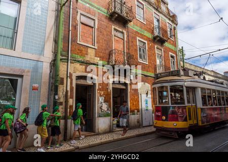 Straßenszene mit Schulkindern und der Straßenbahn Nr. 12 im Baixa-Alfama-Viertel, farbenfrohen Gebäuden, Lebensmittelgeschäften und Fußgängern. Lissabon, Portugal. Stockfoto