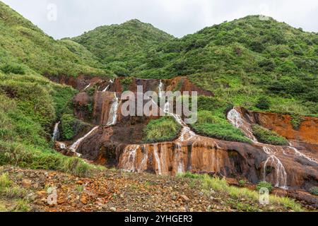 Ein kleiner Wasserfall am Straßenrand in Juifen, Taiwan, mit üppigem grünem Gras und kontrastierenden orange/braunen Felsen, Wasser, das in weißen Nieten überfließt. Stockfoto