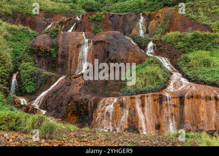 Ein kleiner Wasserfall am Straßenrand in Juifen, Taiwan, mit üppigem grünem Gras und kontrastierenden orange/braunen Felsen, Wasser, das in weißen Nieten überfließt. Stockfoto