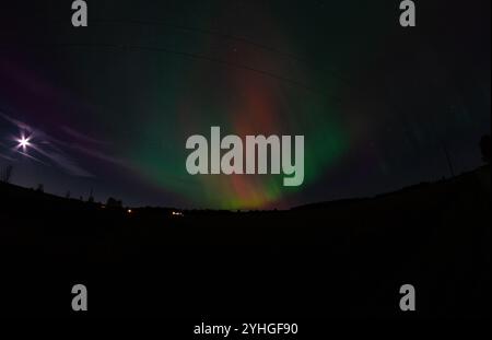 Die aurora Borealis erleuchtet den Nachthimmel über einer ruhigen Farm, mit Stromleitungen, die sich über die Landschaft erstrecken und der Vollmond einen weichen gl erzeugt Stockfoto