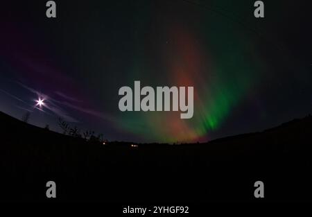 Die aurora borealis zieht über einem Bauernhof über den Himmel, mit Stromleitungen, die sich in die Ferne erstrecken und der Vollmond ein heiteres Licht über den Bauernhof wirft Stockfoto