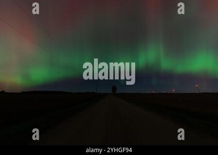 Die aurora borealis erleuchtet den Himmel über einer Landstraße, mit einem einsamen Baum am Straßenrand und Windmühlen, die in der Ferne sichtbar sind, Kreatin Stockfoto