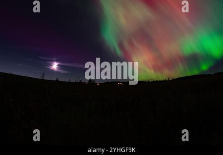 Die aurora Borealis erleuchtet den Himmel über einer ruhigen Farm, mit Stromleitungen über die offenen Felder und Vollmond für eine sanfte, Stockfoto