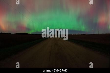 Die aurora Borealis erleuchtet den Himmel über einer friedlichen Landstraße, mit einem einzigen Baum am Straßenrand und Windmühlen im fernen Horizont Stockfoto