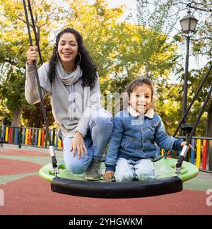 Glückliche hispanische Mutter und Sohn sitzen auf einer Spinnenschaukel auf einem Spielplatz Stockfoto