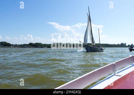 Ein traditionelles Segelschiff gleitet über das ruhige Wasser des Saaler Bodden unter einem hellblauen Himmel mit einem Geländer im Vordergrund. Stockfoto