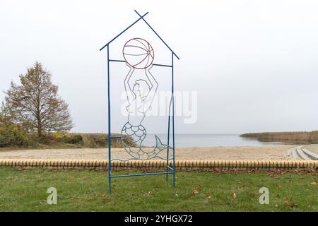 Eine dekorative Metallskulptur einer Meerjungfrau, die einen Ball hält, vor der ruhigen Kulisse von Stettiner Haff an einem bewölkten Novembertag. Stockfoto