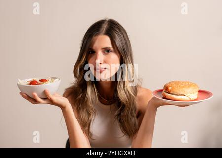 Junge Frau mit gesundem Salat und Hamburger, die das Dilemma bei der Auswahl von Lebensmitteln zeigt, gesunde versus ungesunde Ernährung Lebensstil Konzept Stockfoto
