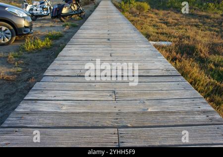 Ein verwinkelter Holzweg führt bei Sonnenuntergang durch goldenes Gras in der Nähe von geparkten Fahrzeugen am Strand. Stockfoto