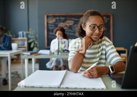 Porträt eines lächelnden afroamerikanischen Teenagers mit Brille und Laptop während des Studiums im Kopierraum der Schule Stockfoto